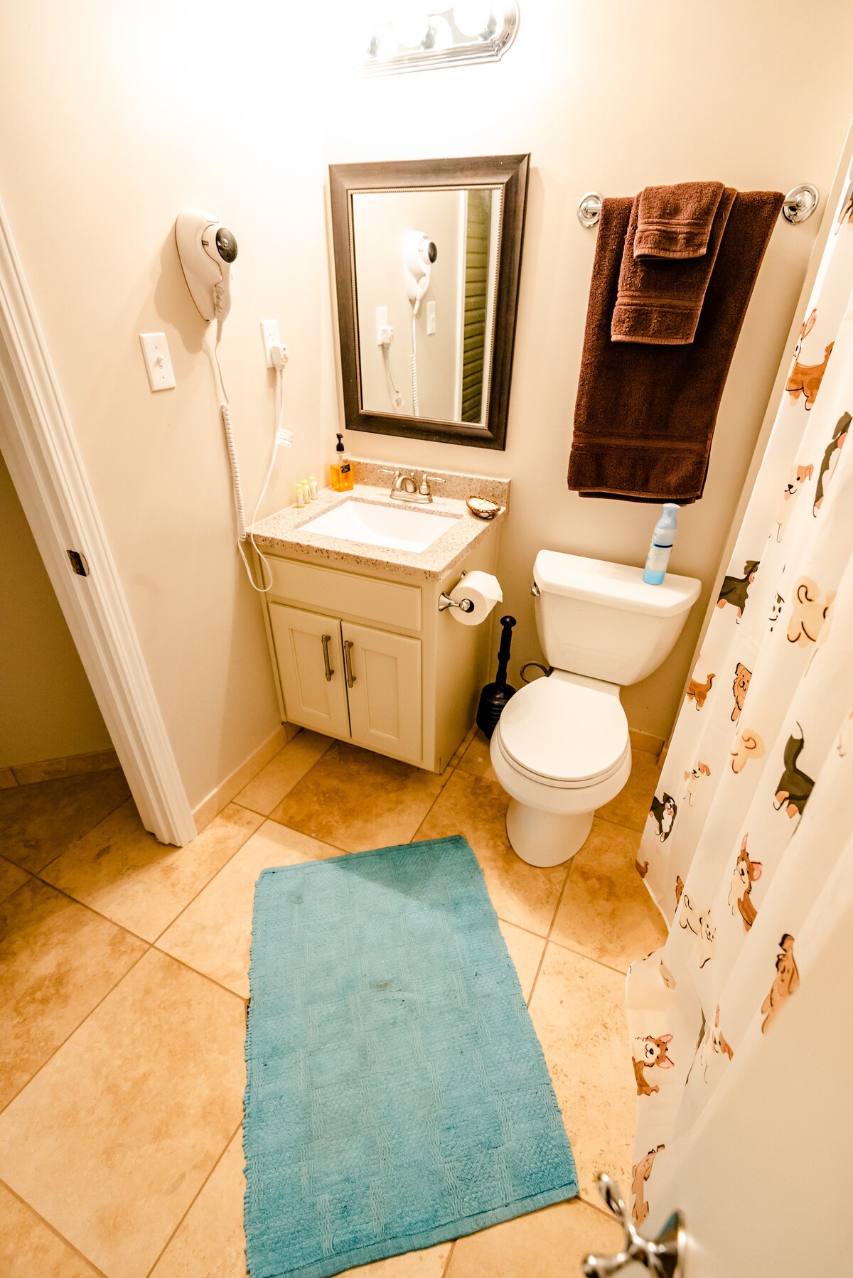 A compact bathroom features a sink with a granite countertop and a mirror above. A toilet is positioned beside the sink. A shower curtain with a playful design adds character, while a turquoise bath mat complements the neutral tile flooring.
