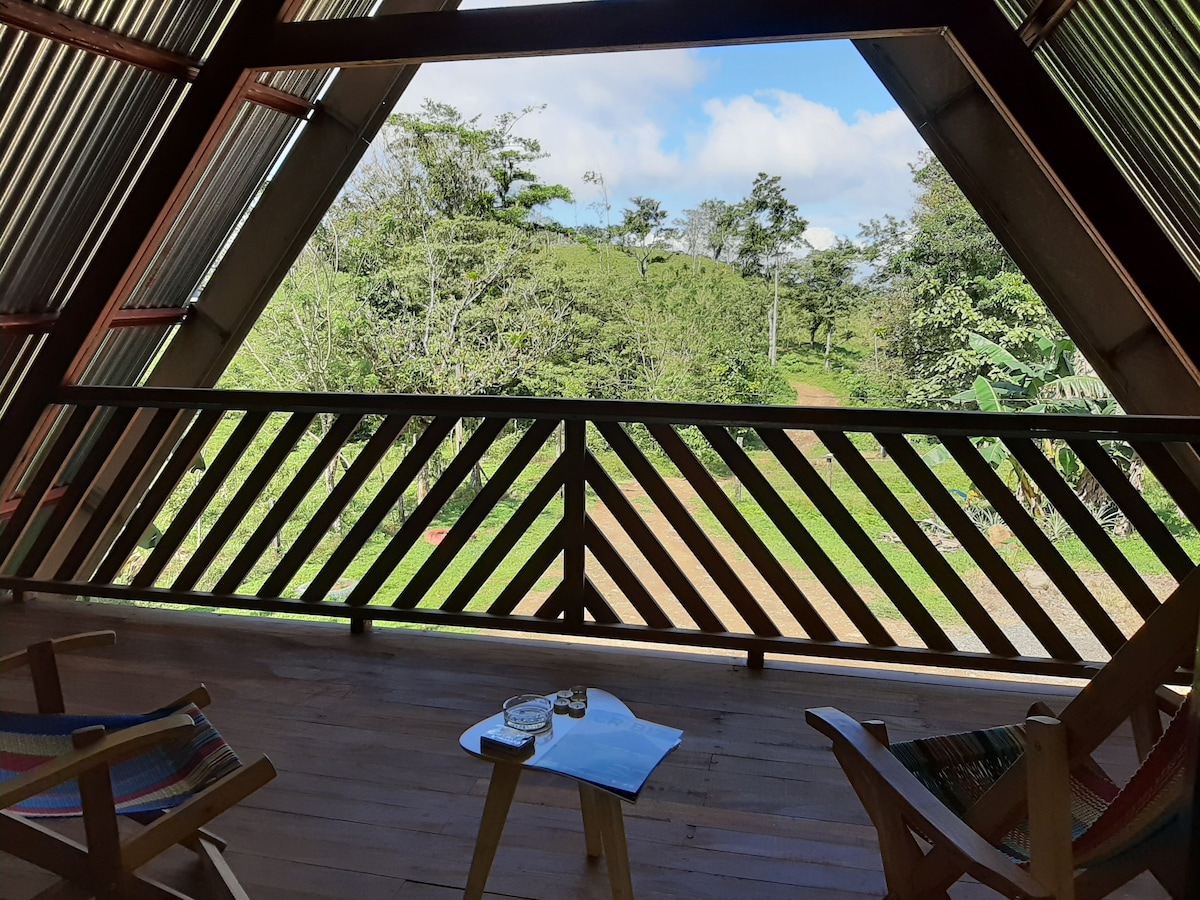 A wooden deck features two colorful chairs, providing a comfortable space to relax and enjoy the natural surroundings. Lush greenery is visible in the background, enhancing the serene atmosphere. The structure's triangular shape and open railing allow ample light and fresh air to enter.