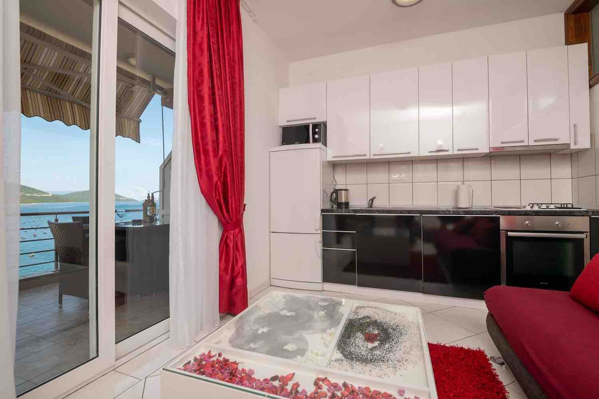 A modern kitchen area is equipped with white cabinetry and black appliances, including a microwave and an oven. A large window overlooks the sea, framed by light-colored curtains. A red sofa complements the space, while a decorative table features a floral design.
