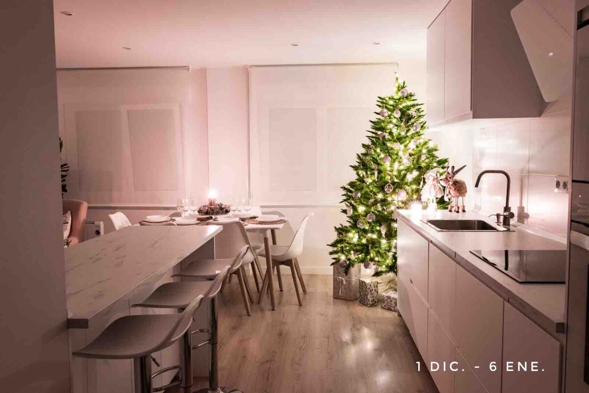 A modern kitchen with a central island and three tall stools is shown. A dining area is visible with a table and white chairs. A decorated Christmas tree stands beside the dining area, providing a festive touch, while soft lighting creates a warm atmosphere.