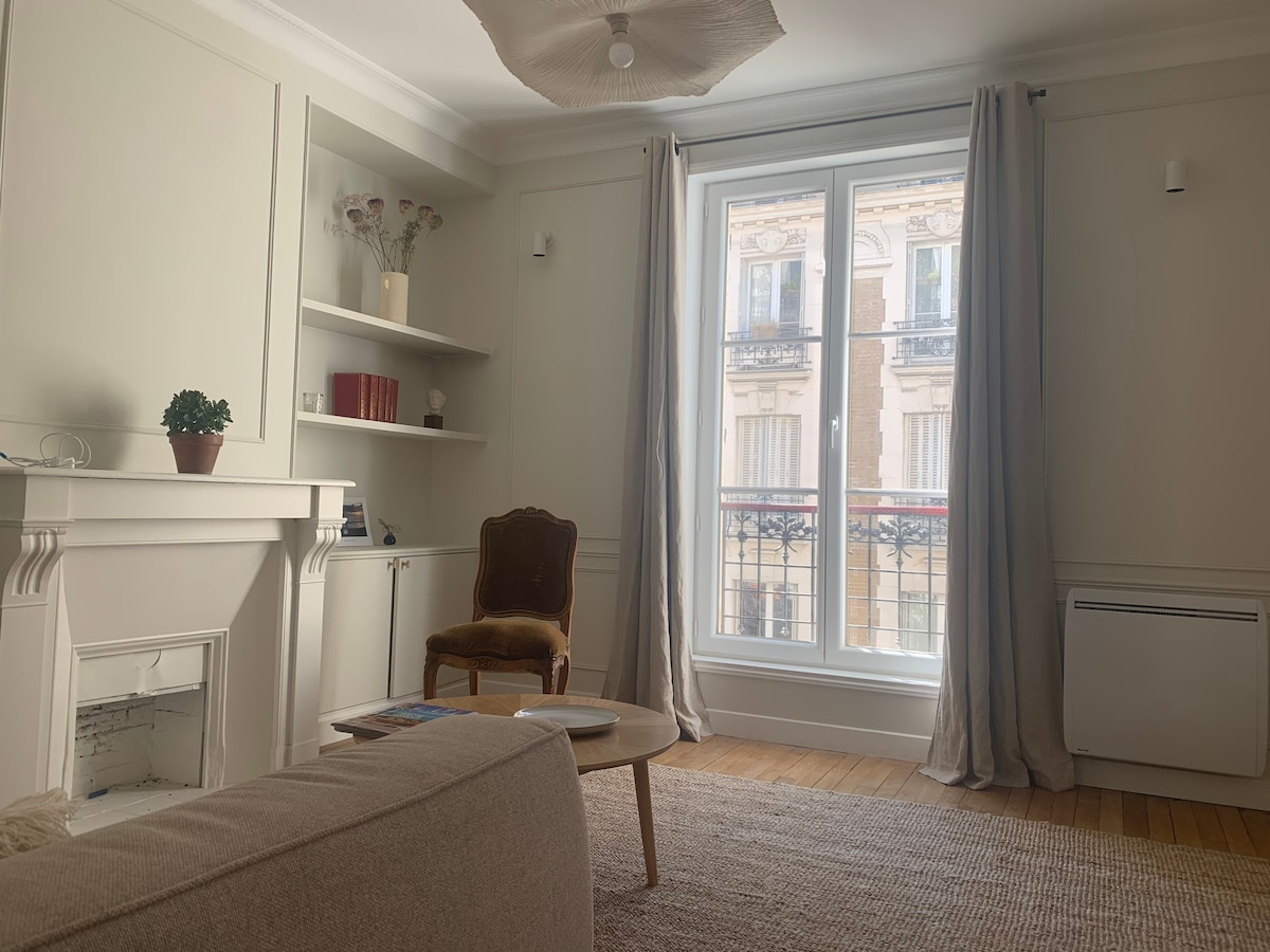 A light-filled living area features a comfortable sofa and a stylish armchair beside a small coffee table. A wall-mounted shelf displays decorative items, while large windows with soft curtains offer views of the lively street outside. Warm wooden flooring enhances the inviting atmosphere.