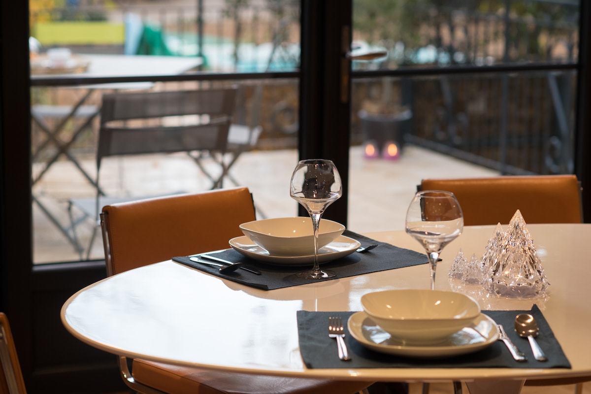 A dining table is set with two bowls and two wine glasses, positioned on black placemats. Soft natural light enters through large glass doors, revealing a view of an outdoor terrace with a bistro set in the background.