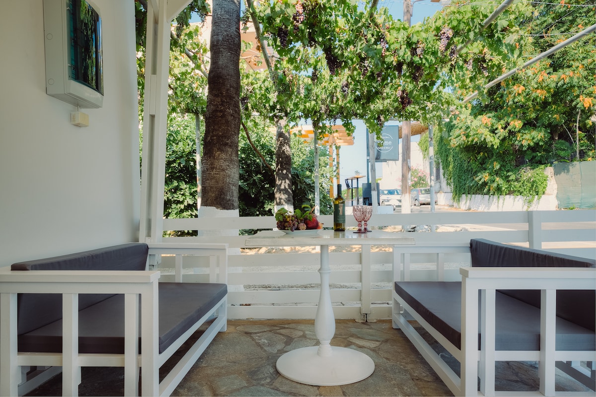 A private patio with a stone floor features white wooden seating arranged around a central table. Lush greenery and hanging vines create a natural canopy above, while a friendly atmosphere is enhanced by nearby trees and colorful plants.