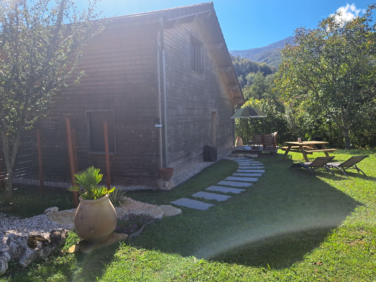 The exterior of a wooden house is surrounded by lush greenery and features a pathway made of stone tiles. Outdoor seating is visible, including loungers and a table with an umbrella. The backdrop includes gently rolling hills and a clear blue sky.
