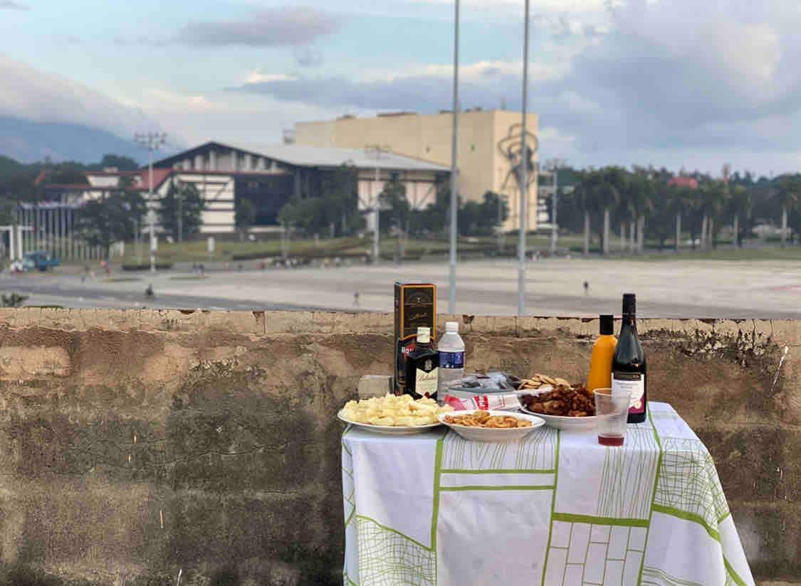 A table is set outdoors with a white tablecloth, displaying a variety of dishes, drinks including red and white wine, and snacks. In the background, a spacious plaza and modern buildings are visible, indicating a lively urban area.
