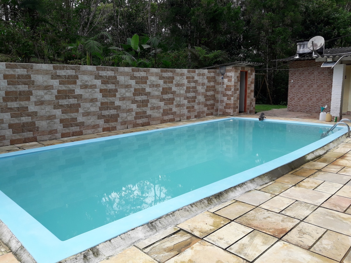 A rectangular swimming pool is surrounded by a tiled patio, featuring light-colored stone tiles. The pool water is clear and calm, with lush greenery visible in the background. A brick structure is partially visible to the right, adding to the serene outdoor setting.