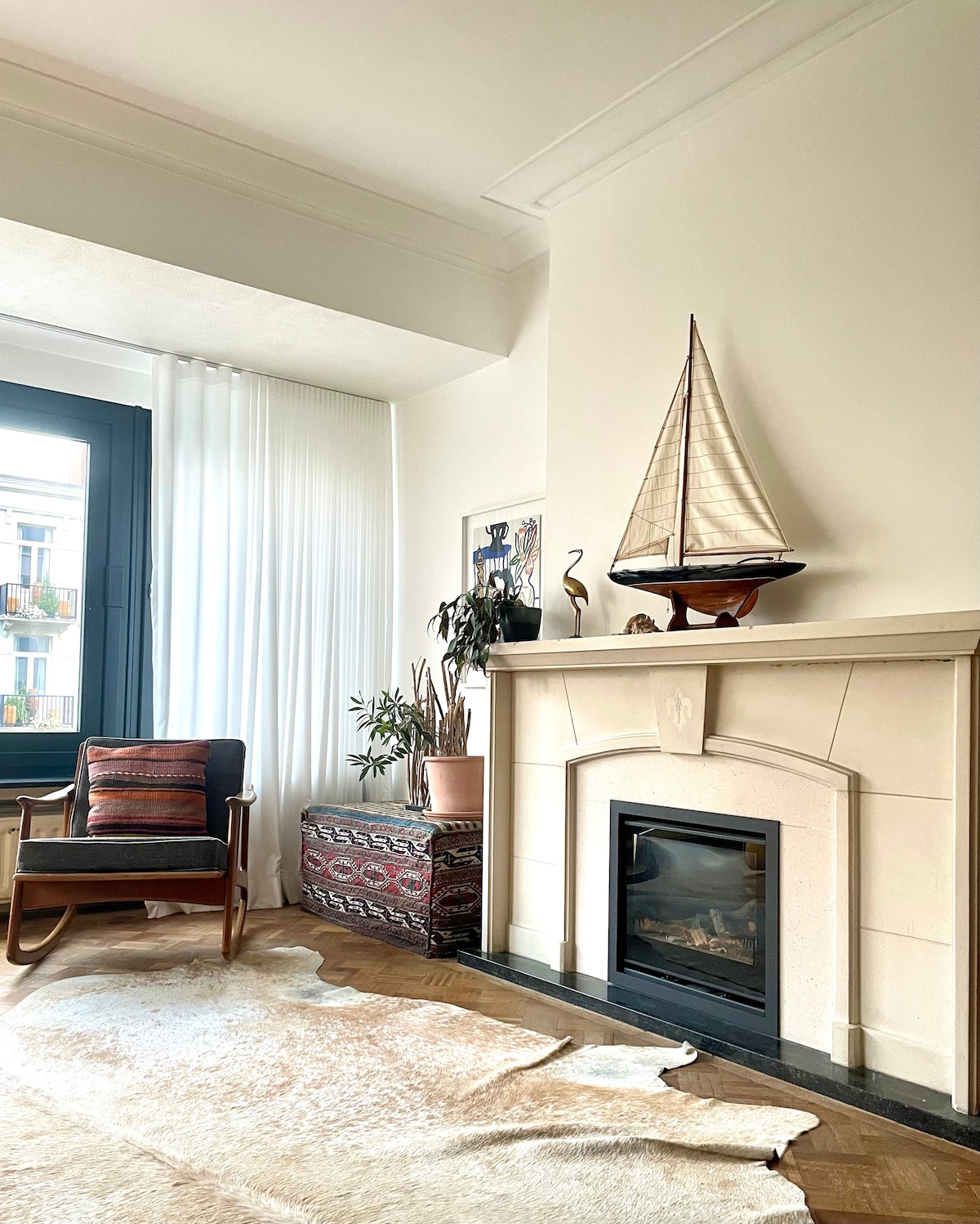 A modern living area is depicted, featuring a stylish chair and an elegant wooden fireplace adorned with a decorative sailboat. Light curtains frame a large window, allowing natural light to brighten the space. A patterned rug complements the warm tones of the decor.