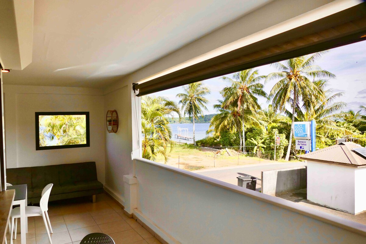 A spacious living area is shown, featuring large windows that provide a view of the surrounding palm trees and a glimpse of the water. A comfortable sofa is positioned to one side, alongside a dining table with chairs.