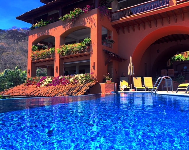 An inviting infinity pool reflects the vibrant blue sky, surrounded by lush greenery. The building features arched balconies adorned with colorful flowers, enhancing its charm. Comfortable lounge chairs are positioned nearby, offering a relaxing space to enjoy the stunning views.