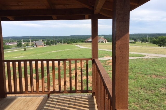 A porch area with wooden railing offers an expansive view of green fields and distant homes. The landscape is open, showcasing a peaceful countryside setting under a clear sky. The wooden floor enhances the natural ambiance of the space.