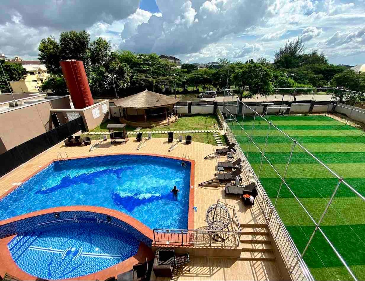 A view of the outdoor pool area is captured, featuring a sparkling blue pool surrounded by wooden decking. Lounge chairs are positioned near the pool, while a shaded gazebo sits nearby. An expansive green football pitch is visible in the background, framed by trees under a partly cloudy sky.
