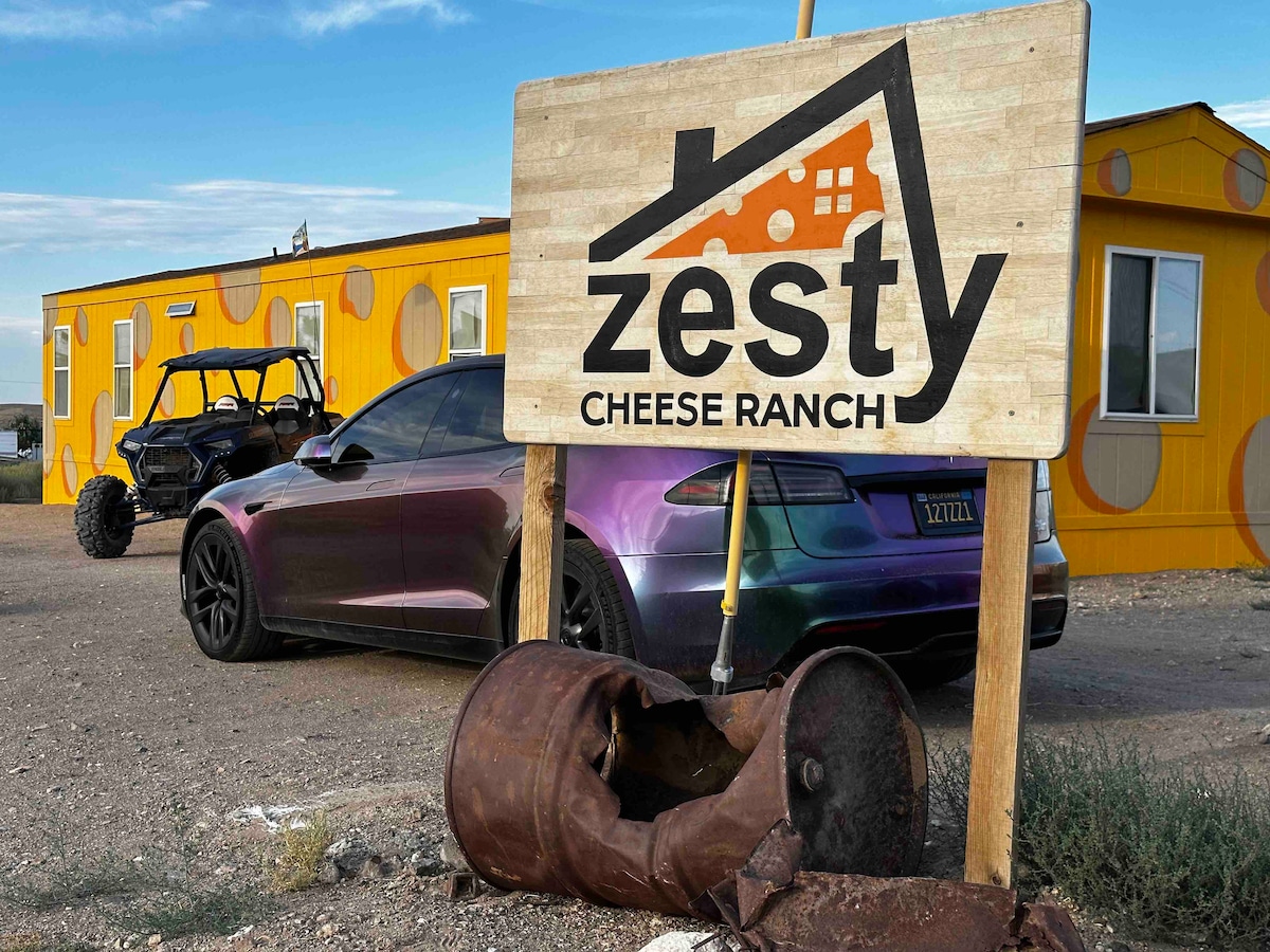 A rustic sign for Zesty Cheese Ranch is positioned in front of a yellow structure, displaying a logo with a house icon. A dark silver car is parked nearby, alongside a utility vehicle. An old barrel rests on the ground, adding a touch of character to the scene.