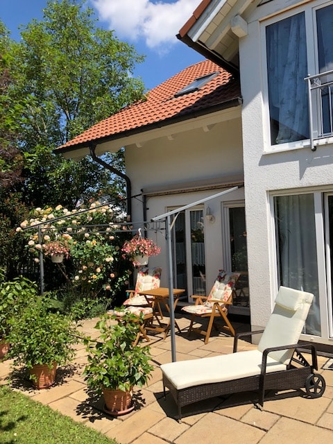 A patio area is displayed, featuring a sun lounger and two wooden chairs with floral cushions. Lush greenery surrounds the space, with blooming plants in terracotta pots and a gazebo adorned with hanging flowers. The home's exterior is showcased, with large windows allowing natural light.