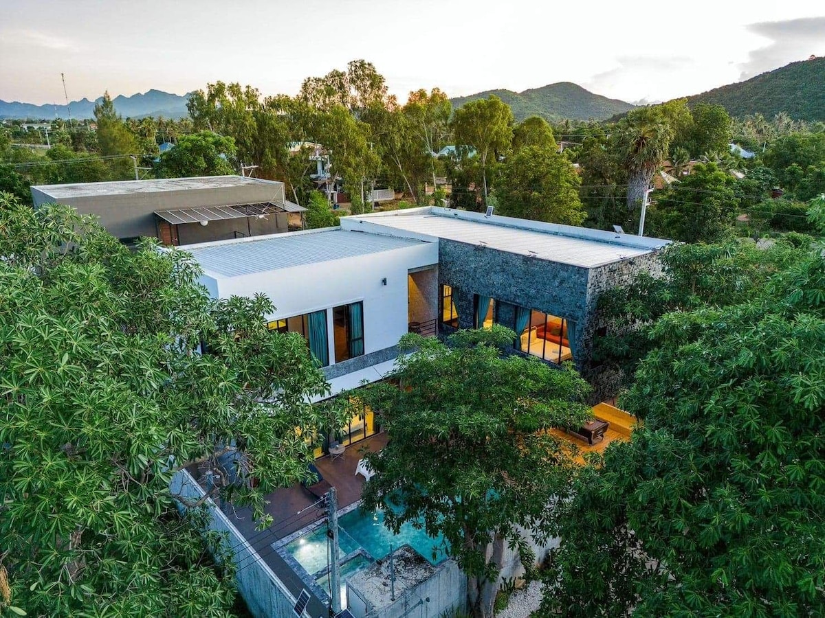 An aerial view of Villa Rocka is presented, showcasing the modern architectural design surrounded by lush greenery. The structure features expansive windows that invite natural light, with a well-kept pool and bubbling Jacuzzi visible in the backyard, inviting relaxation and leisure.