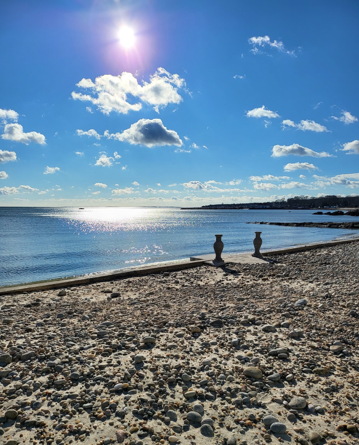 The tranquil shoreline reveals a pebbled beach alongside calm waters, reflecting sunlight. A clear blue sky, dotted with clouds, creates a serene atmosphere. Silhouettes of two vases can be seen on the shore, adding a touch of artistic charm to the natural setting.