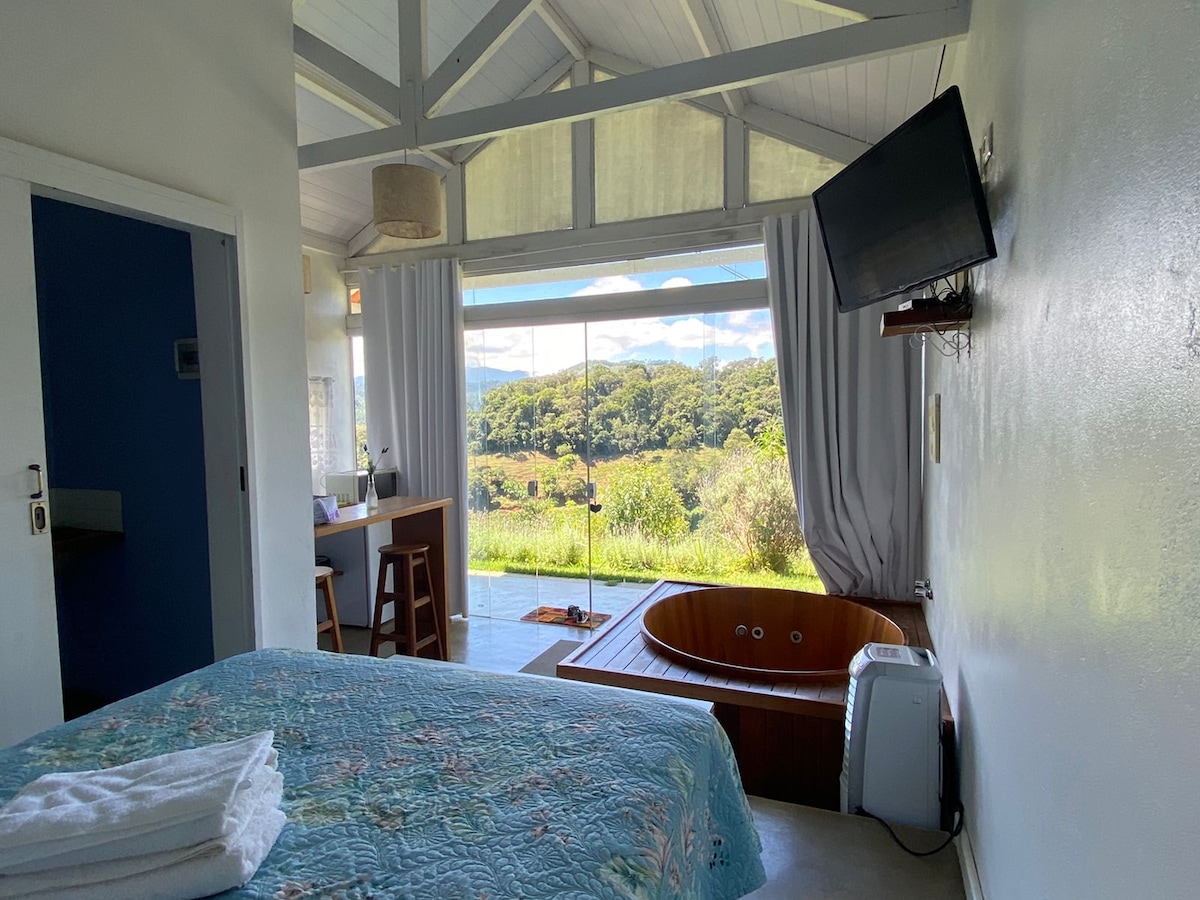 The inviting interior features a spacious bed with light blue linens and a cozy quilt. A wooden hot tub is positioned near a window, which provides scenic views of lush greenery. Soft natural light filters through sheer curtains, enhancing the serene atmosphere.