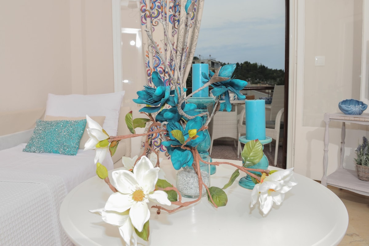 A coffee table is adorned with a decorative arrangement featuring turquoise and white flowers along with tall blue candles in glass holders. In the background, a balcony view is visible, framed by elegant curtains that complement the decor.