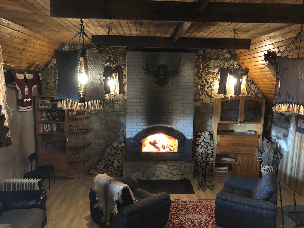 A rustic living area features a central stone fireplace with a glowing fire. Wooden beams line the ceiling, and warm lighting is provided by hanging fixtures. Two comfortable armchairs are positioned near the fireplace, while stacked firewood decorates a corner. Natural textures and elements are present throughout.