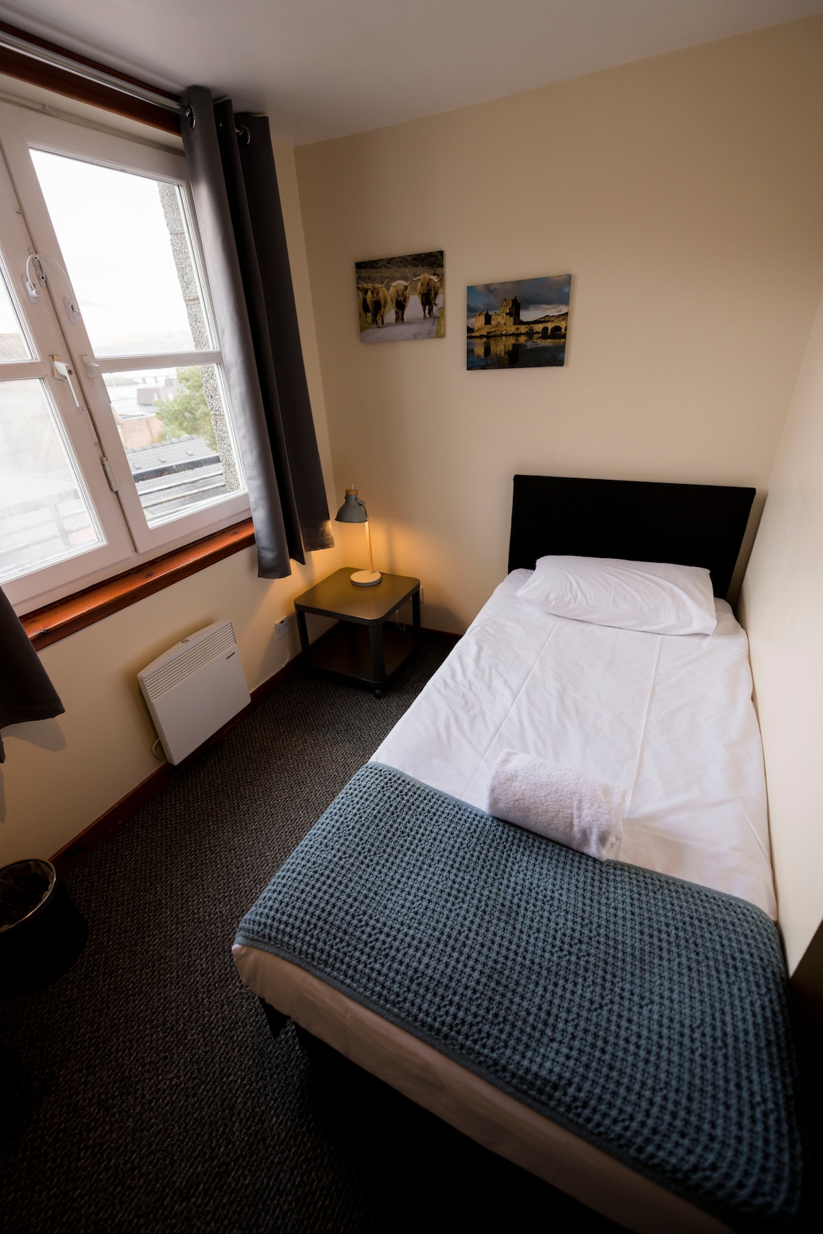 A compact room features a single bed with a light blue blanket. A small bedside table with a lamp is positioned next to the bed. Two framed photographs are displayed on the wall above the bed, and natural light enters through a window covered by gray curtains.
