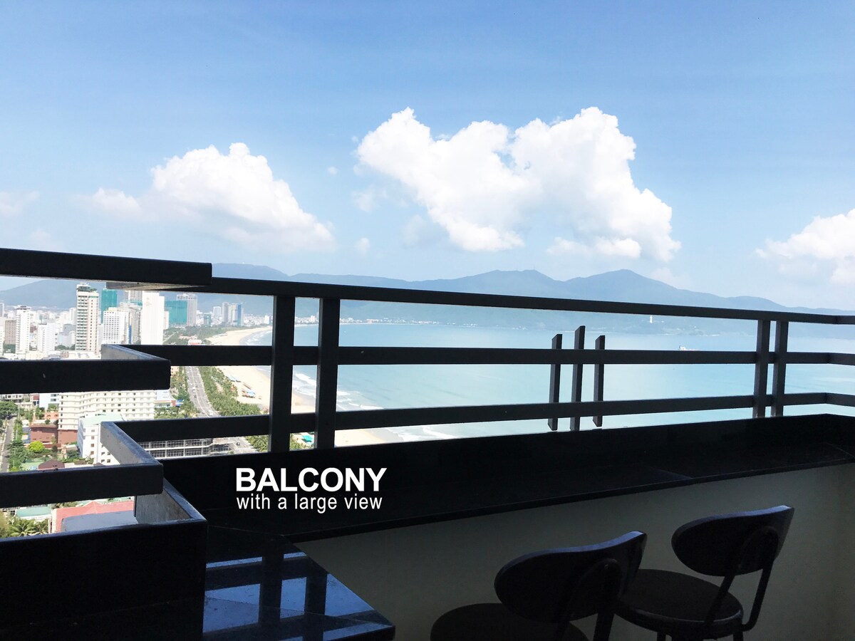 The balcony offers a spacious view of the sea and surrounding cityscape. High chairs are positioned for comfort, allowing guests to relax while enjoying the scenery. A clothesline is also visible, providing practical space for hanging laundry.