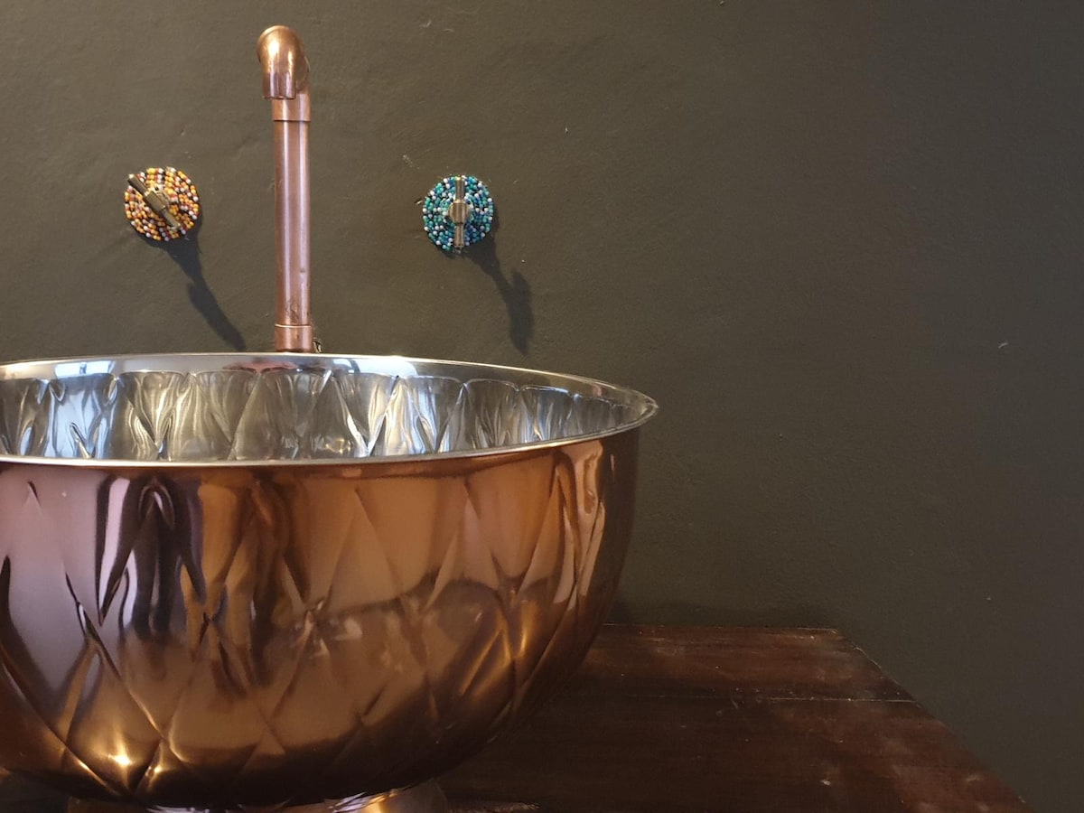 A polished copper sink is showcased against a dark wall. The sink's intricate diamond pattern reflects light, while two decorative wall fixtures, embellished in vibrant colors, are positioned above. The rustic wooden countertop enhances the overall aesthetic of the space.