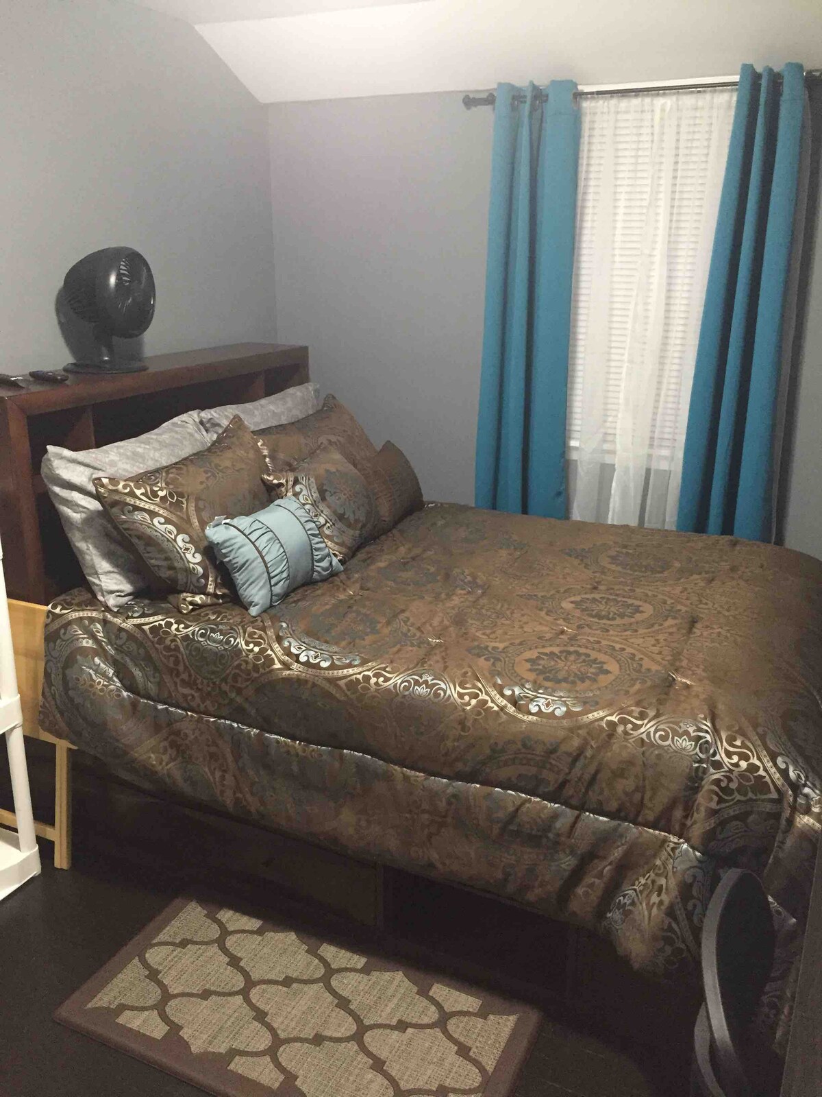 A cozy bedroom features a comfortable bed with a decorative brown and silver bedding set. Blue accent pillows complement the look. The space is illuminated by natural light from a nearby window, draped with white sheer and blue curtains. A small fan and a rug add practicality to the room.