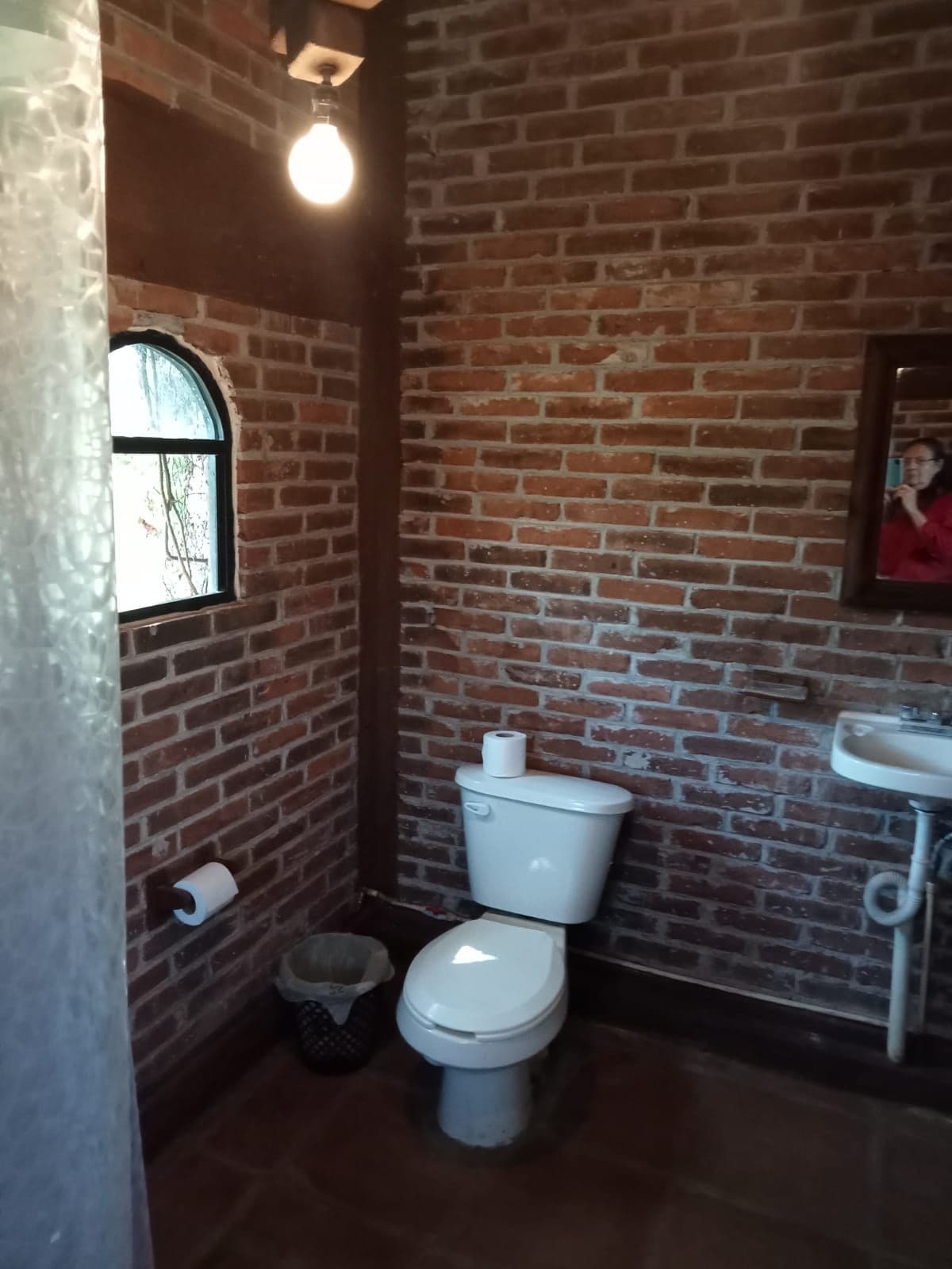 The bathroom features a rustic design with exposed brick walls, complemented by a small window allowing natural light. A toilet and a sink are positioned conveniently, with a towel rack and wastebasket nearby. A shower area is separated by a translucent curtain.