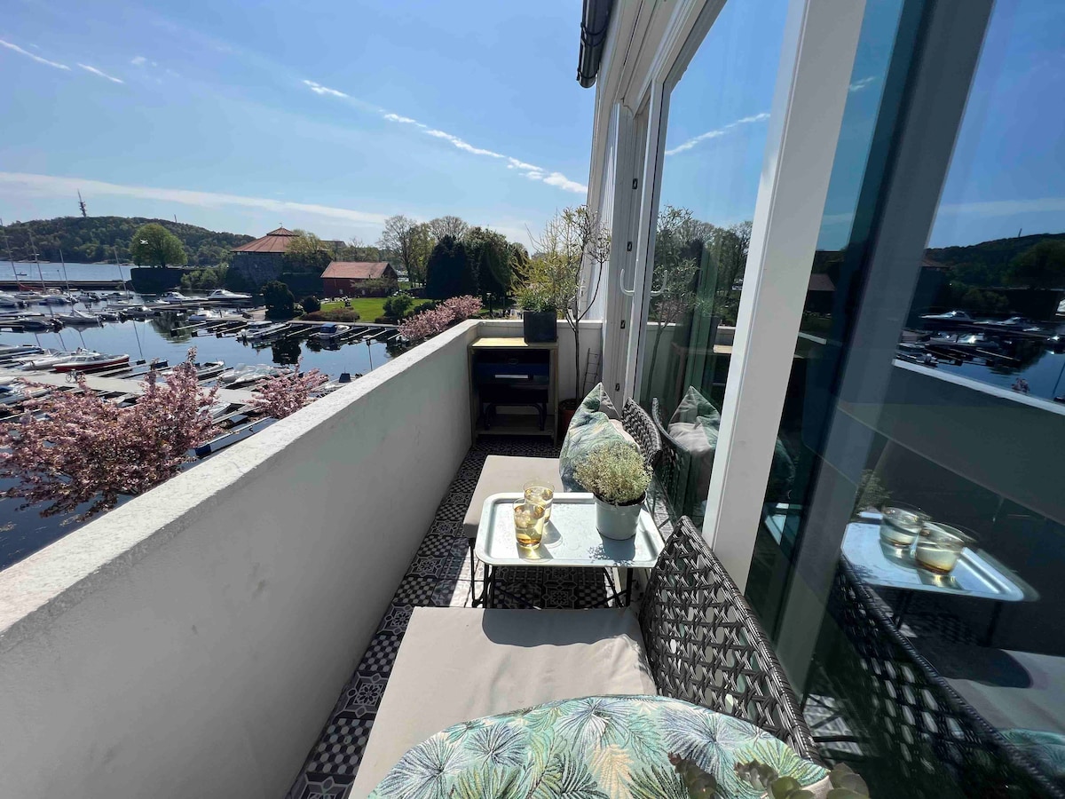 A balcony area is presented with two cushioned lounge seats facing a serene view of the marina and surrounding greenery. A small table positioned nearby holds decorative plants and glassware, while large windows reflect the bright sunlight and vibrant outdoor scene.