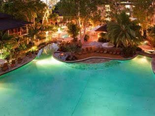 An expansive swimming pool is surrounded by lush greenery, with soft lighting creating a serene atmosphere. Comfortable lounge chairs are positioned along the water's edge, inviting relaxation. In the background, additional palm trees and resort amenities are visible, enhancing the inviting resort setting.