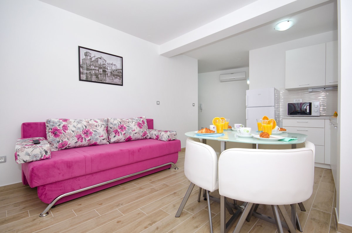 A modern living area features a vibrant pink sofa adorned with floral patterns. A round dining table is set with dishes and glasses, contributing to a cozy atmosphere. The space includes a kitchenette with essential appliances, while light-colored walls enhance the airy feel.