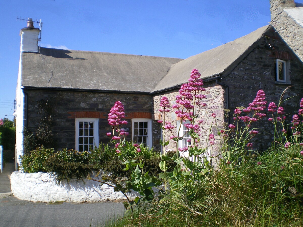 Yewdale Cottage is framed by vibrant pink flowers, showcasing its charming stone facade. The thatched roof and classic windows add to its character, while the surrounding greenery complements the inviting atmosphere of this traditional home.