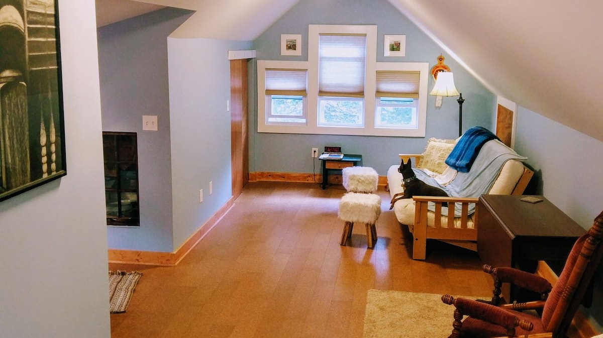 A spacious loft area features a futon couch with a light blanket draped over it. Two soft stools and a side table complement the cozy seating arrangement. A rocking chair offers additional comfort, while three windows provide natural light, framed by light-colored shades.