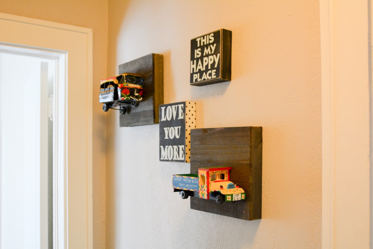 A warm corner features decorative wall art, including small signs and toy trucks mounted on wooden plaques. The signs convey messages of happiness and love, contributing to a welcoming ambiance. A light-colored wall complements the darker wooden accents.