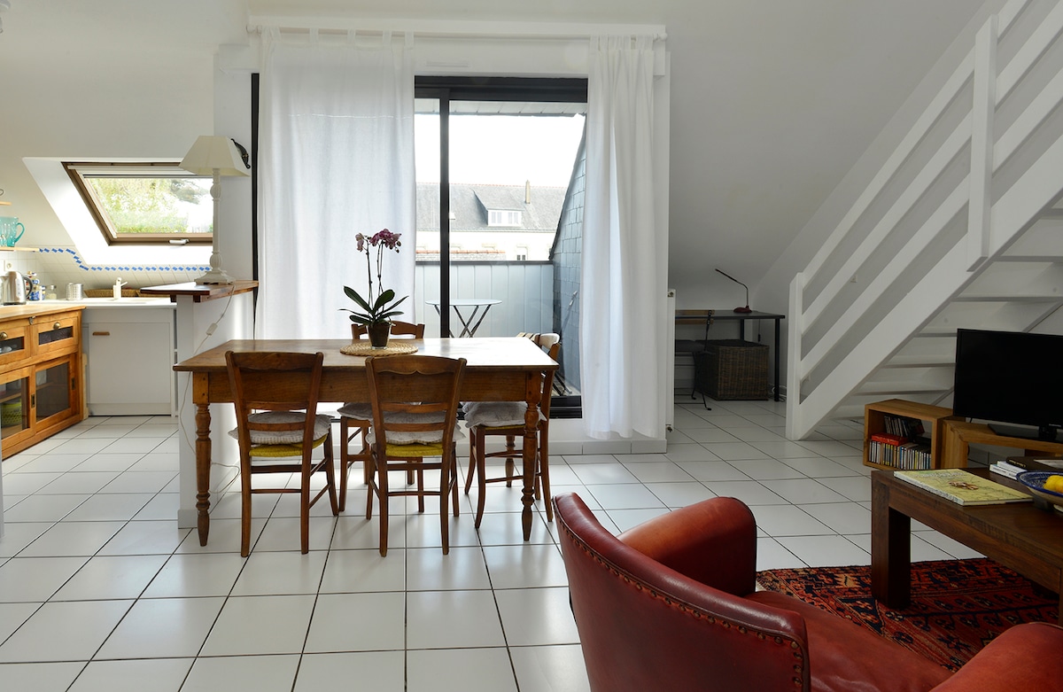 A bright living space is highlighted by large windows with sheer white curtains, allowing natural light to fill the room. A wooden dining table with several chairs is situated at the center, accompanied by a comfortable armchair and a small television on a stand.