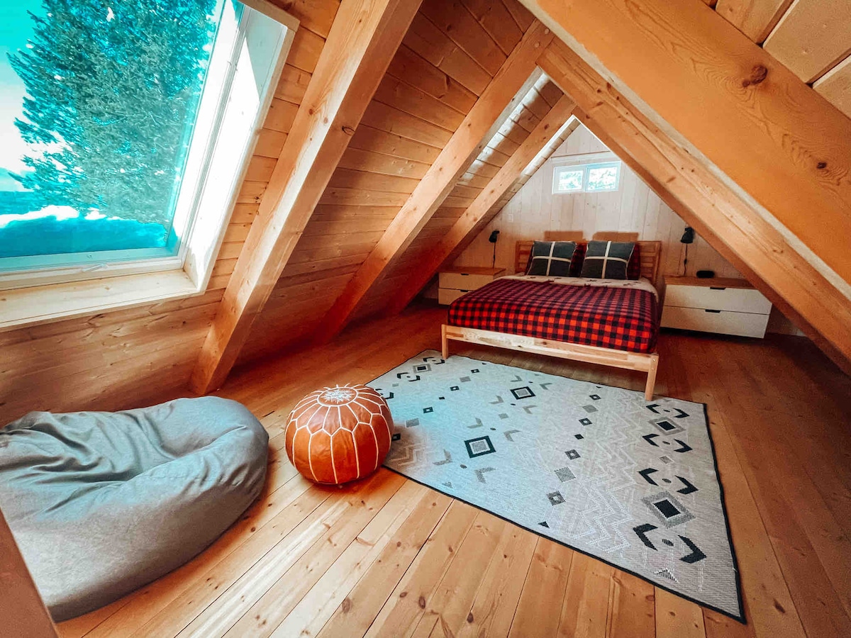 A warm attic bedroom features a spacious bed dressed in a plaid blanket. A cozy bean bag sits beside a patterned rug on the wooden floor. Large windows illuminate the room, allowing natural light to highlight the exposed wooden beams and ceiling.