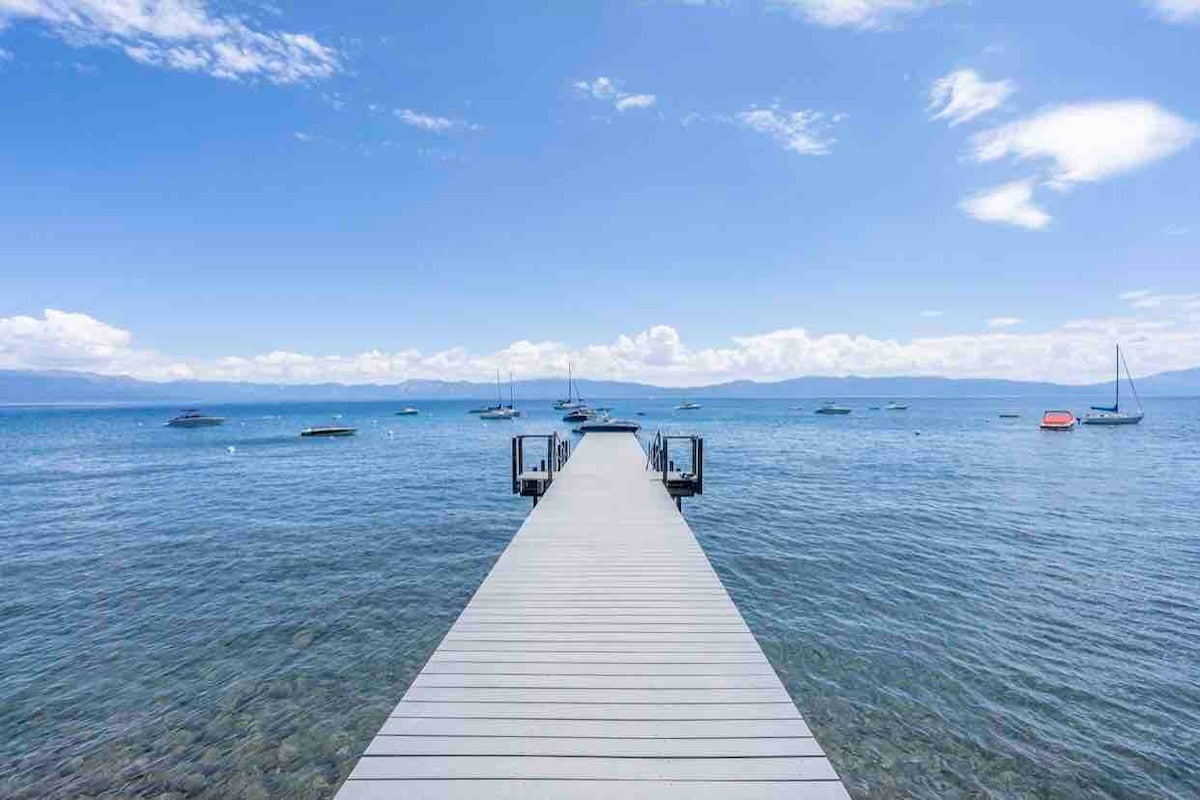 A wooden dock extends into the clear blue waters of Lake Tahoe, surrounded by distant mountain views under a sunny sky. Various boats float on the surface, contributing to a serene lakeside atmosphere.