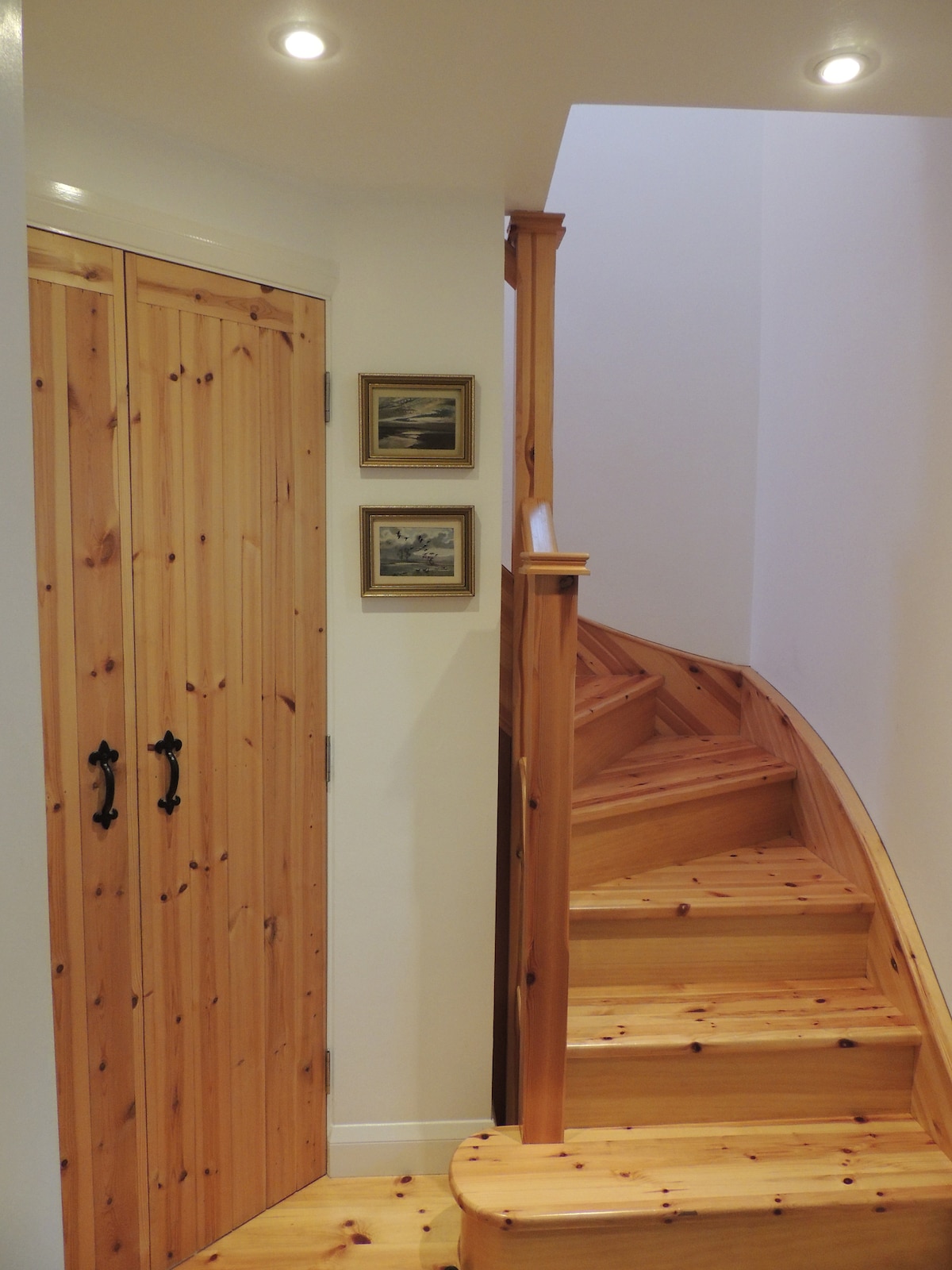 A wooden staircase curves upwards, leading to the next level of the cottage. The walls are painted in a light color, complemented by recessed lighting. A set of closed wooden doors and two framed pictures are positioned neatly to the left.