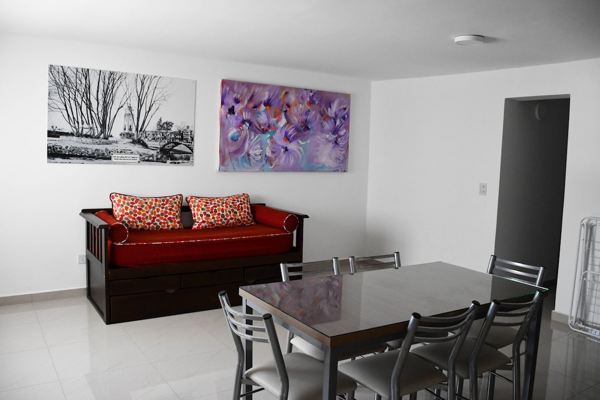 A dining area is presented with a rectangular table surrounded by six metal chairs. A wooden sofa with vibrant cushions is positioned against the wall. Two large wall art pieces add color to the space, with a bright floral painting and a black and white landscape photograph.