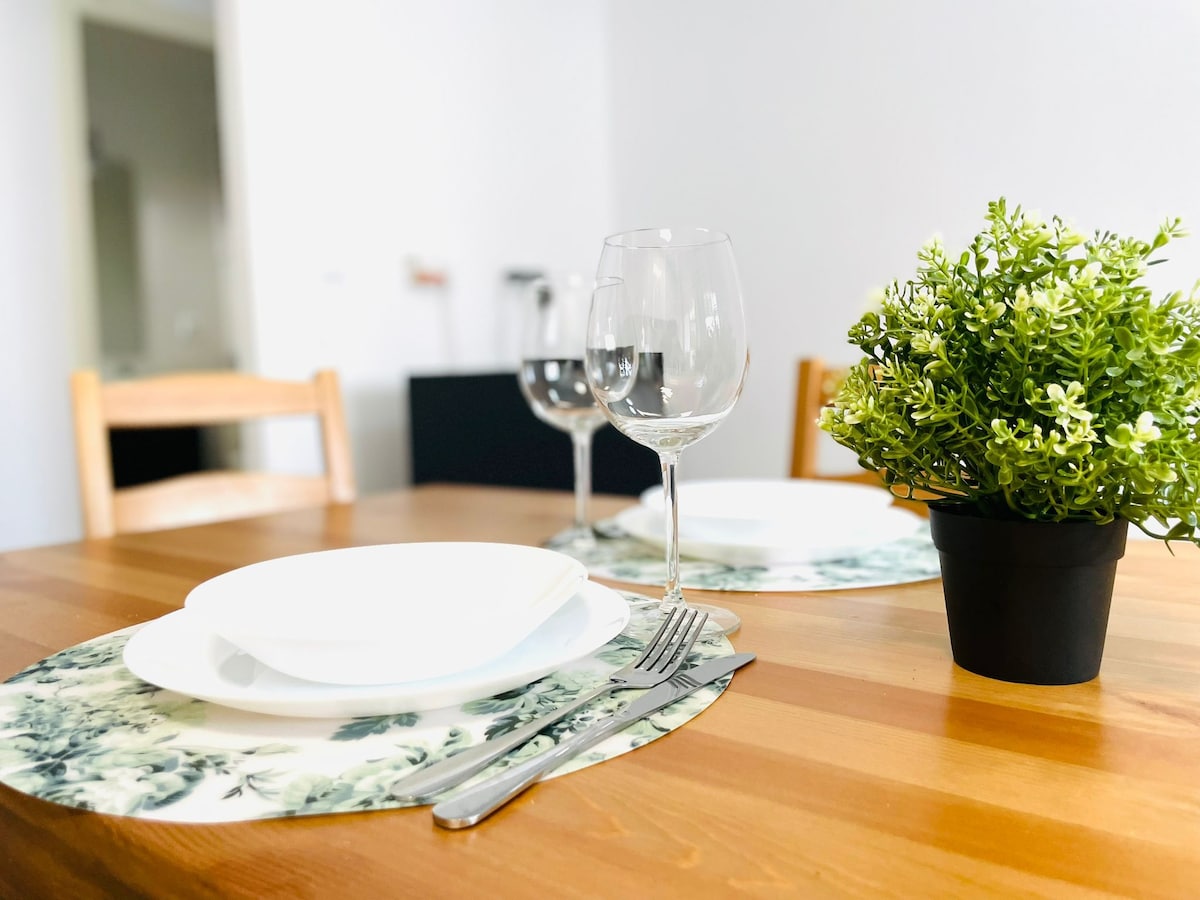 A dining table set for two is arranged with white plates and silverware, complemented by glasses ready for use. A small potted plant adds a touch of greenery, while the wooden table features a natural finish, enhancing its inviting appearance.