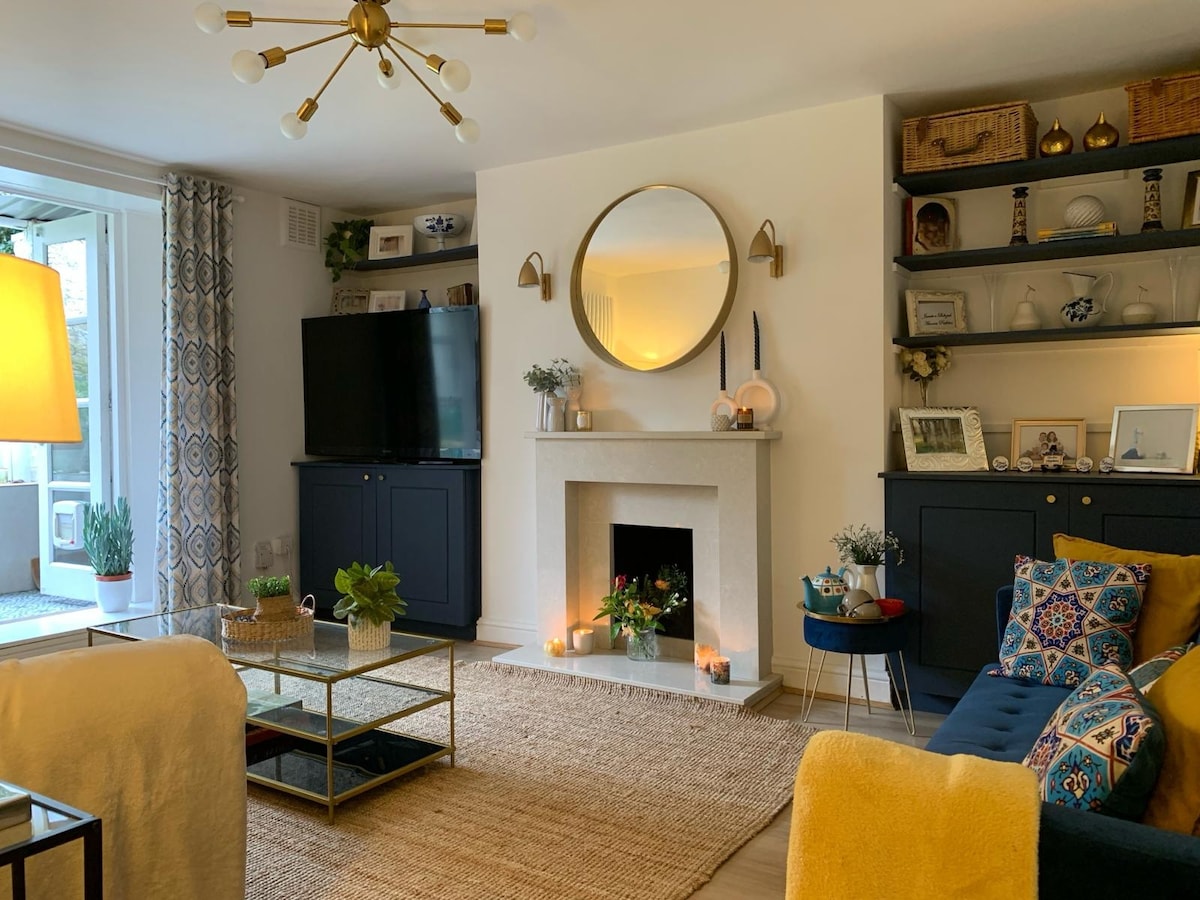 A cozy lounge area is highlighted by a stylish blue sofa and a glass coffee table atop a woven rug. A flat-screen television is mounted on a light-colored wall, complemented by a decorative fireplace. Natural light enters through glass doors, creating a bright and inviting atmosphere.