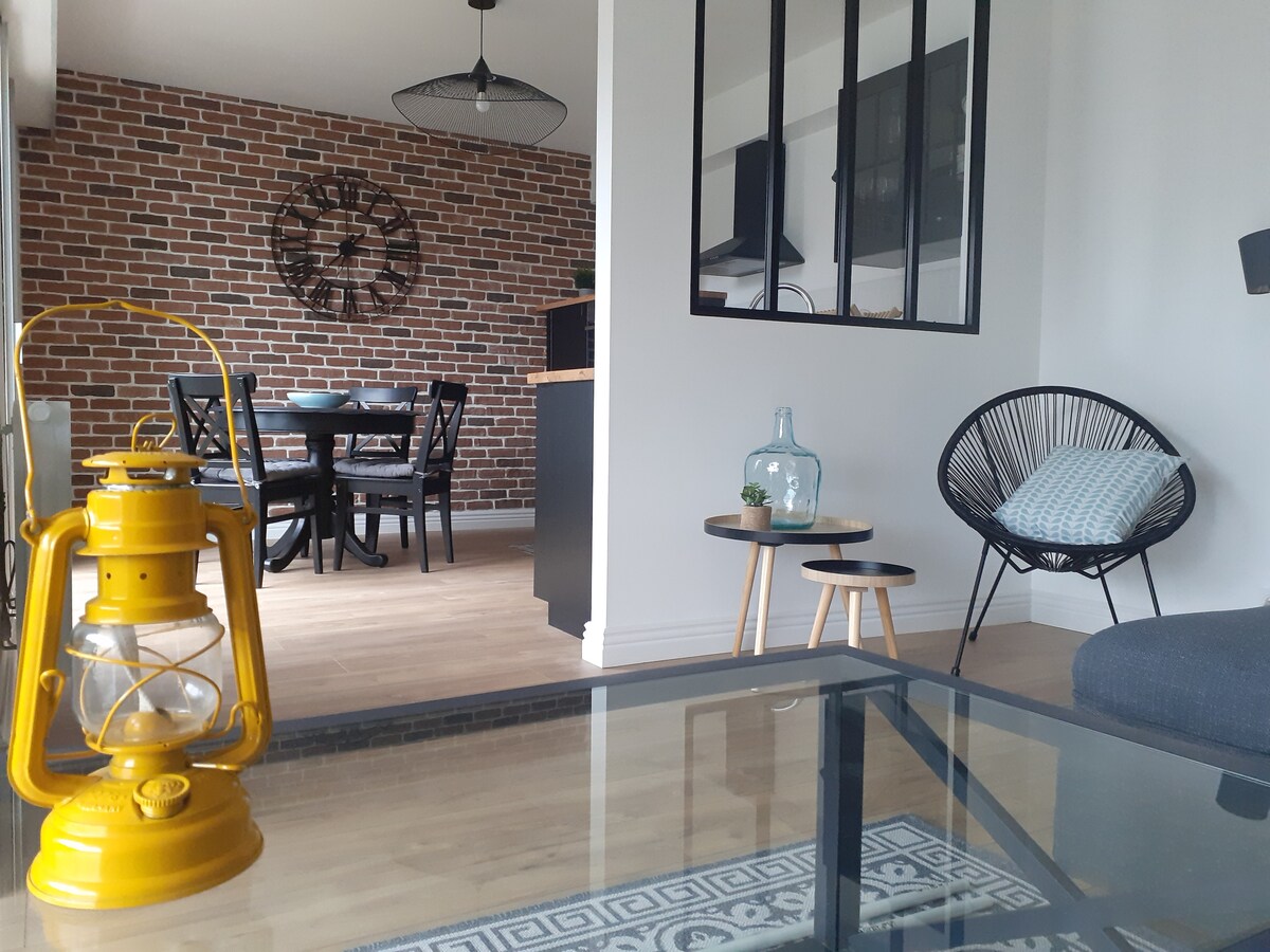 An inviting living space features a glass coffee table and a vintage yellow lantern. A cozy seating area with a dark sofa overlooks a dining area with a round table and black chairs. The backdrop is enhanced by a brick wall, adding character to the room.