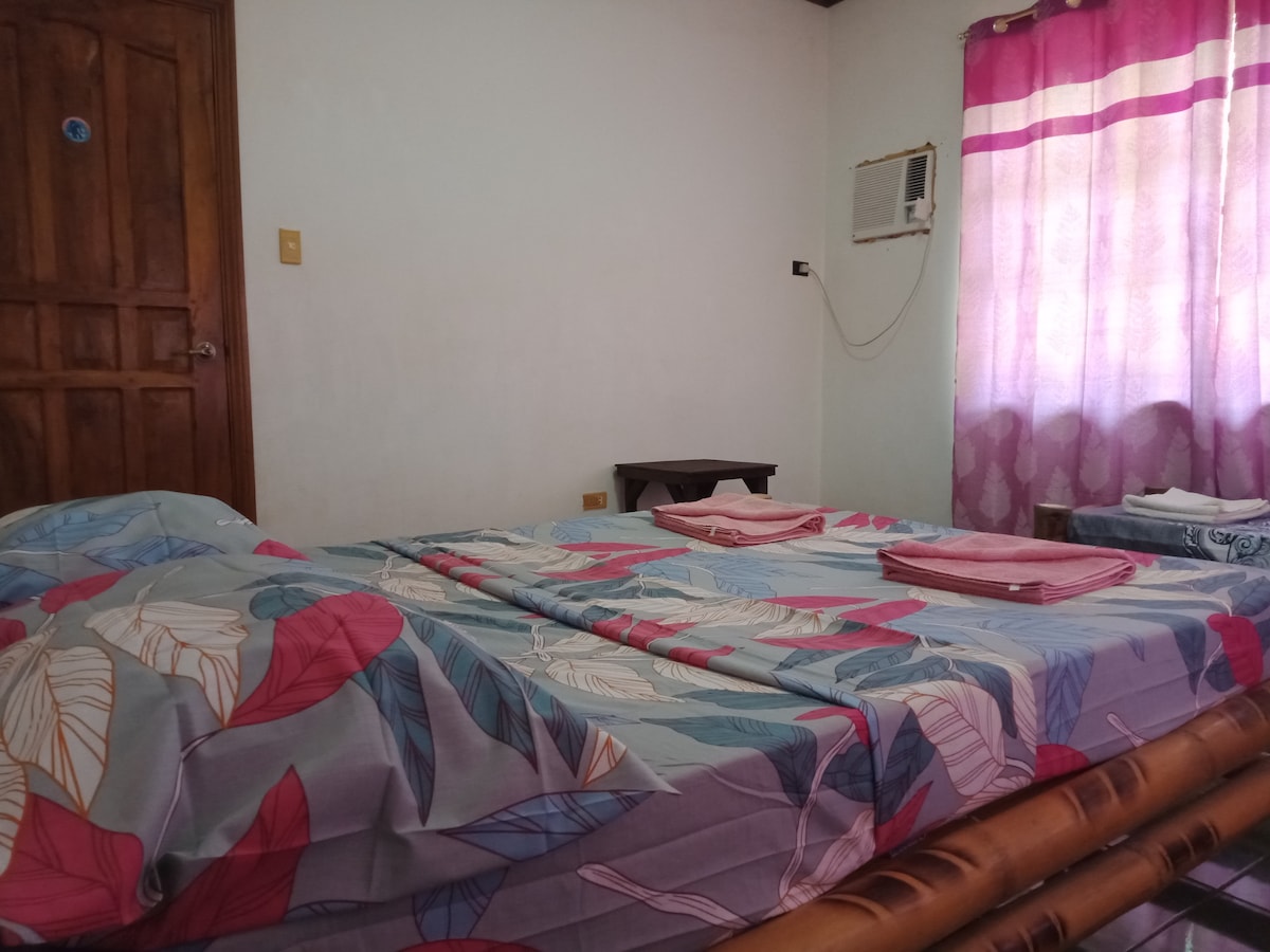 The room features a large bed adorned with a floral patterned blanket. A small wooden table is positioned to the side, while light filters through a window dressed in semi-transparent curtains. An air conditioning unit is visible, indicating current ventilation conditions.