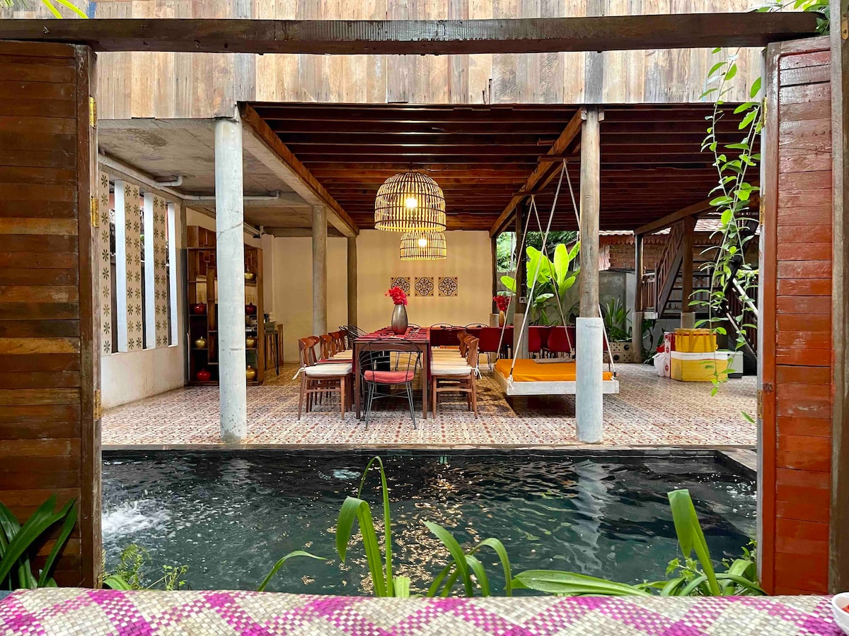 An inviting open space features a private plunge pool with surrounding greenery and vibrant cement tiles. A dining area with a large table is complemented by comfortable seating. Natural light enters through spacious openings, enhancing the airy atmosphere within the traditional structure.