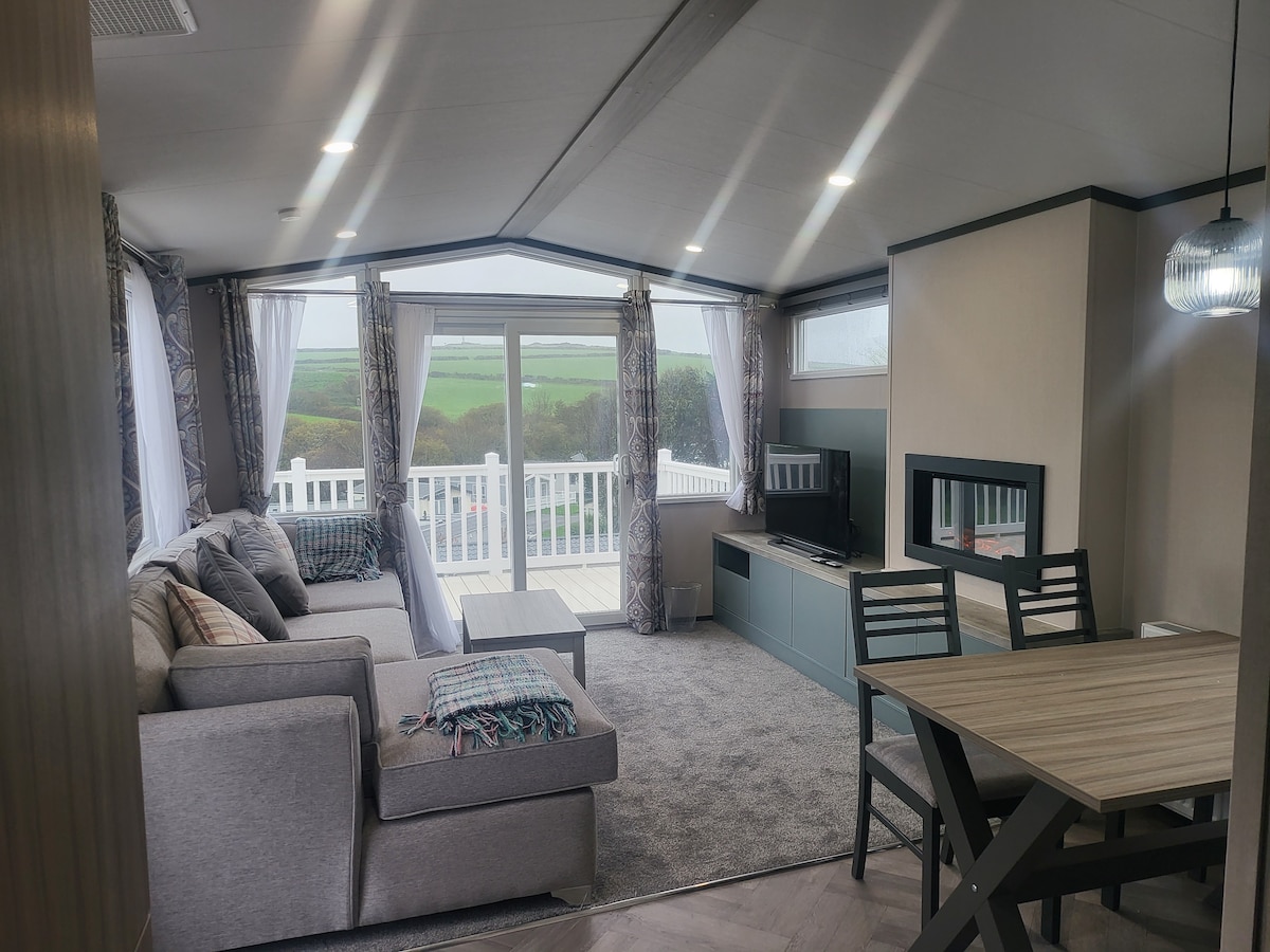 A spacious living area is highlighted by large glass doors leading to a deck, allowing natural light to fill the space. Comfortable seating is arranged around a modern entertainment unit, complemented by a dining table and chairs. Scenic valley views are visible through the windows.