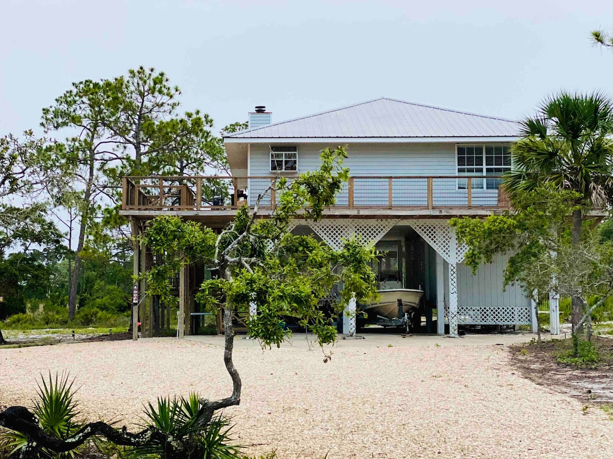 The exterior of the property features a two-story structure with a light-colored facade and a metal roof. A spacious deck extends from the upper floor, while a boat is visible underneath. Surrounding vegetation includes pine trees and low shrubs, creating a natural setting.