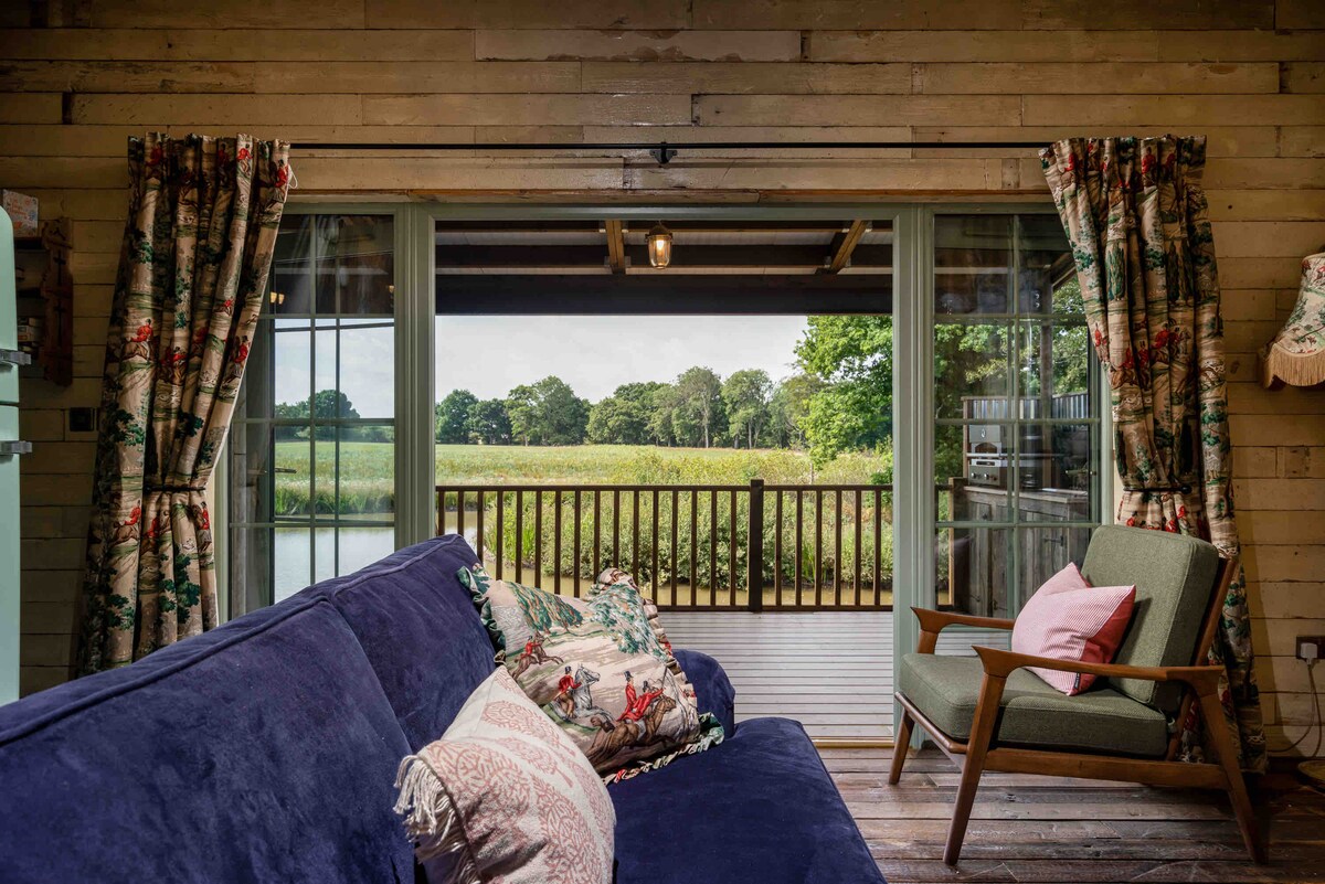 A comfortable seating area is visible, featuring a plush blue sofa adorned with patterned cushions. Large glass doors open to a balcony overlooking lush greenery and a serene lake, while floral-patterned curtains frame the entrance, adding a touch of rustic charm.