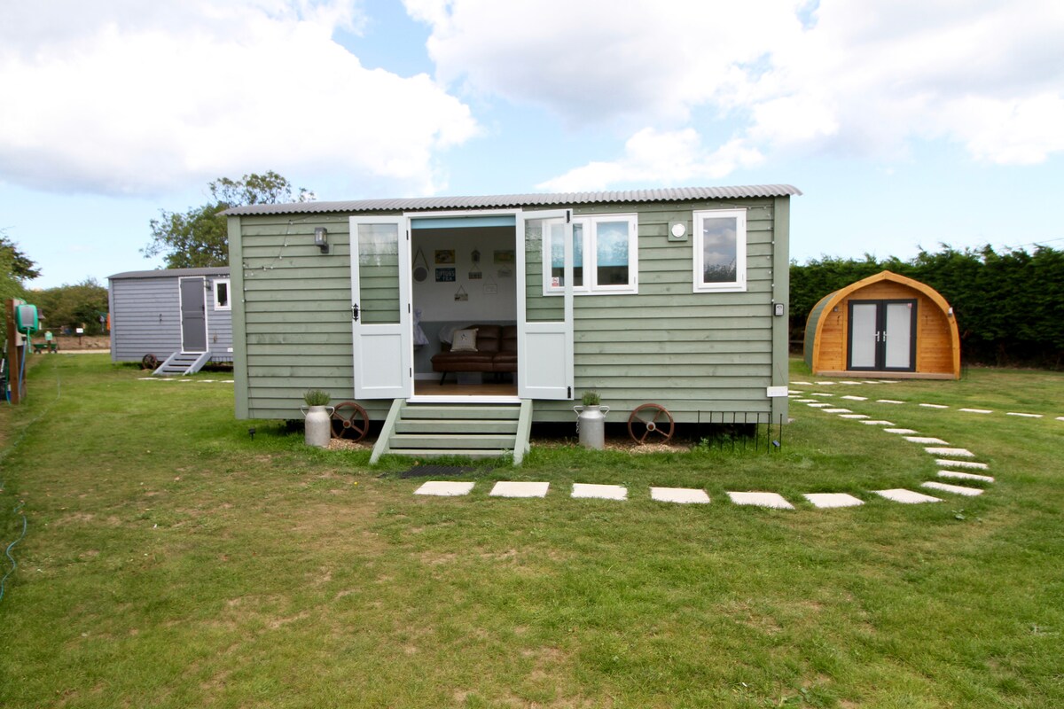 A green shepherd's hut is viewed from the exterior, featuring French doors that open onto a grassy area. The path is made of stepping stones, leading towards a wooden arch-shaped pod and other huts. Lush greenery surrounds the setting, creating a peaceful ambiance.