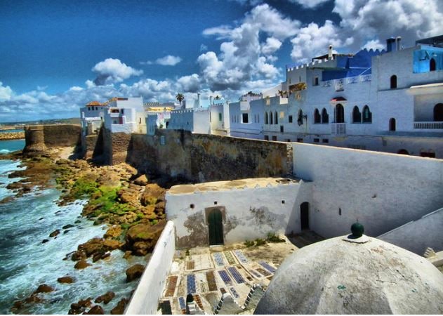 A coastal view features a whitewashed fortress wall against a backdrop of blue skies and scattered clouds. The rocky shoreline meets the sea, while traditional buildings with blue accents line the top of the wall, creating a harmonious blend with the natural landscape.
