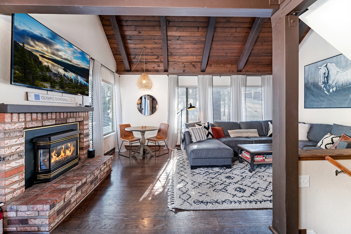 The living area features a modern sectional sofa, circular dining table, and a decorative fireplace surrounded by a brick mantel. Large windows invite natural light, and a flat-screen TV is mounted on the wooden-beamed ceiling. Decorative art adds a touch of style to the space.
