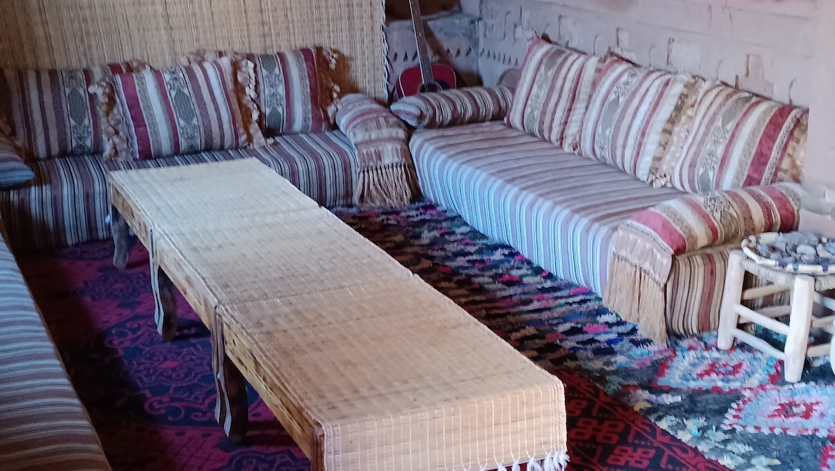 The lounging area features low, woven furniture with patterned cushions. A light-colored, textured table is centered on a colorful rug, while traditional wall decorations add to the ambiance of the space.